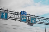 Multiple blue traffic signs are installed on a metal overhead structure. These signs include directional arrows pointing straight, right, and downward, with accompanying text. The background consists of a cloudy sky with some patches of blue visible.