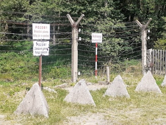 A barbed wire fence with several warning signs in Czech and German languages, surrounded by triangular concrete barriers. The signs display warnings about a border zone, and the area is backed by dense greenery, including trees and grass.
