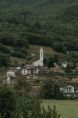 A picturesque village nestled at the base of a lush, forested hillside. The village features a variety of small houses with different roof colors, dominated by a tall, white church tower topped with a green roof. The landscape shows terraced fields and dense greenery surrounding the settlement, giving it a serene and peaceful atmosphere.