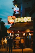 A street scene at night features brightly lit neon signs advertising a sex shop, pub, and brasserie. The signs are colorful and prominent against the dark evening backdrop, attracting attention. People are walking along the sidewalk, adding to the urban night atmosphere.