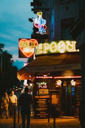 A street scene at night features brightly lit neon signs advertising a sex shop, pub, and brasserie. The signs are colorful and prominent against the dark evening backdrop, attracting attention. People are walking along the sidewalk, adding to the urban night atmosphere.