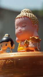 The image features three small, cartoon-style figurines with serene expressions. In the center, a figure resembling the Buddha is depicted with closed eyes and a gentle smile, adorned with golden hair. On the left, a smaller figure with black hair holds a blue object, possibly a small animal. On the right, another character holds a golden accessory and wears a crown with a symbol. They are placed on a surface engraved with intricate patterns and Chinese characters.