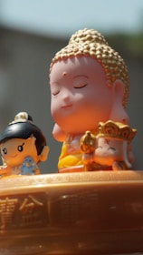 The image features three small, cartoon-style figurines with serene expressions. In the center, a figure resembling the Buddha is depicted with closed eyes and a gentle smile, adorned with golden hair. On the left, a smaller figure with black hair holds a blue object, possibly a small animal. On the right, another character holds a golden accessory and wears a crown with a symbol. They are placed on a surface engraved with intricate patterns and Chinese characters.