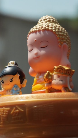 The image features three small, cartoon-style figurines with serene expressions. In the center, a figure resembling the Buddha is depicted with closed eyes and a gentle smile, adorned with golden hair. On the left, a smaller figure with black hair holds a blue object, possibly a small animal. On the right, another character holds a golden accessory and wears a crown with a symbol. They are placed on a surface engraved with intricate patterns and Chinese characters.