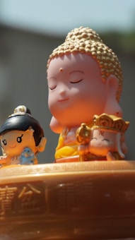 The image features three small, cartoon-style figurines with serene expressions. In the center, a figure resembling the Buddha is depicted with closed eyes and a gentle smile, adorned with golden hair. On the left, a smaller figure with black hair holds a blue object, possibly a small animal. On the right, another character holds a golden accessory and wears a crown with a symbol. They are placed on a surface engraved with intricate patterns and Chinese characters.