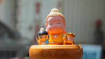 A small, serene Buddha figurine with closed eyes is seated in the center, accompanied by two smaller cartoon-like figures on either side. The setting suggests peacefulness and harmony, with a hint of playfulness.