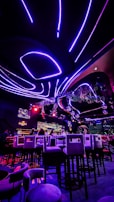 A vibrant interior of a nightclub or bar with futuristic elements. Neon purple and red lights illuminate the space, creating a visually striking atmosphere. High bar stools are arranged in front of a counter where people are seated. The ceiling features curved, glowing lines with hanging decorations, and there are screens displaying visuals.