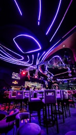 A vibrant interior of a nightclub or bar with futuristic elements. Neon purple and red lights illuminate the space, creating a visually striking atmosphere. High bar stools are arranged in front of a counter where people are seated. The ceiling features curved, glowing lines with hanging decorations, and there are screens displaying visuals.