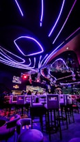 A vibrant interior of a nightclub or bar with futuristic elements. Neon purple and red lights illuminate the space, creating a visually striking atmosphere. High bar stools are arranged in front of a counter where people are seated. The ceiling features curved, glowing lines with hanging decorations, and there are screens displaying visuals.