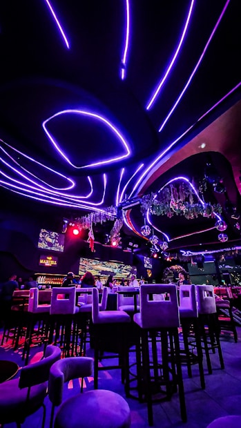 A vibrant interior of a nightclub or bar with futuristic elements. Neon purple and red lights illuminate the space, creating a visually striking atmosphere. High bar stools are arranged in front of a counter where people are seated. The ceiling features curved, glowing lines with hanging decorations, and there are screens displaying visuals.
