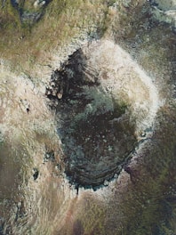 Aerial view of a large crater-like formation on rocky, uneven terrain. The surrounding area is covered with scattered rocks and sparse vegetation. The center appears darker, possibly indicating deeper or varied material composition.