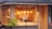 A cozy restaurant with large windows, featuring warm lighting from hanging pendant lamps. The interior shows wooden walls and decor, with patrons sitting inside at a table. Outside, the building is adorned with a small palm plant and a tiled roof.