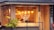 A cozy restaurant with large windows, featuring warm lighting from hanging pendant lamps. The interior shows wooden walls and decor, with patrons sitting inside at a table. Outside, the building is adorned with a small palm plant and a tiled roof.