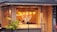 A cozy restaurant with large windows, featuring warm lighting from hanging pendant lamps. The interior shows wooden walls and decor, with patrons sitting inside at a table. Outside, the building is adorned with a small palm plant and a tiled roof.