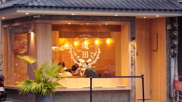 A cozy restaurant with large windows, featuring warm lighting from hanging pendant lamps. The interior shows wooden walls and decor, with patrons sitting inside at a table. Outside, the building is adorned with a small palm plant and a tiled roof.
