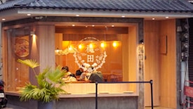 A cozy restaurant with large windows, featuring warm lighting from hanging pendant lamps. The interior shows wooden walls and decor, with patrons sitting inside at a table. Outside, the building is adorned with a small palm plant and a tiled roof.