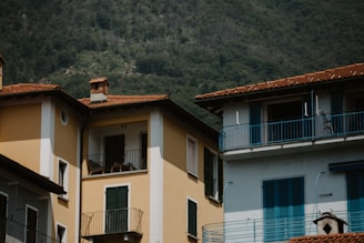 A residential area featuring buildings with multicolored facades and terracotta roofing, set against a backdrop of dense green forest. There are balconies with dark shutters and railings, and the structure reflects a Mediterranean architectural style.