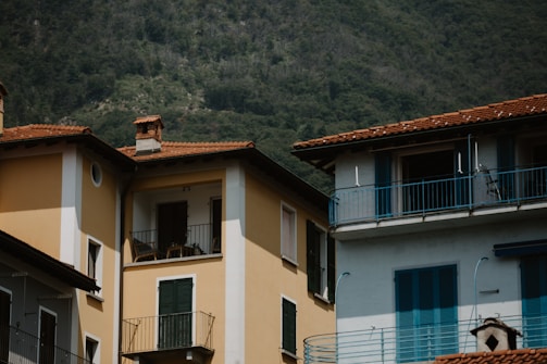 A residential area featuring buildings with multicolored facades and terracotta roofing, set against a backdrop of dense green forest. There are balconies with dark shutters and railings, and the structure reflects a Mediterranean architectural style.