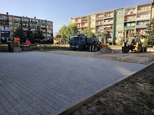 A construction site in an urban setting with a newly paved area in the foreground. In the background, there are various construction vehicles including a dump truck and two backhoes. Residential apartment buildings with multiple balconies are visible behind the construction site. Some trees and playground equipment are also present, adding a touch of greenery and leisure to the scene.
