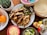 A colorful and inviting spread of food ingredients for making a meal, featuring a plate of grilled chicken strips and sautéed onions and peppers, surrounded by elements like shredded cheese, lime wedges, tortillas, orange fruits, various condiments, and a small cactus plant.