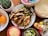 A colorful and inviting spread of food ingredients for making a meal, featuring a plate of grilled chicken strips and sautéed onions and peppers, surrounded by elements like shredded cheese, lime wedges, tortillas, orange fruits, various condiments, and a small cactus plant.