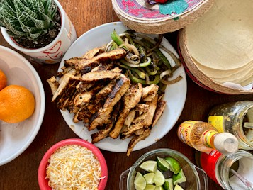 A colorful and inviting spread of food ingredients for making a meal, featuring a plate of grilled chicken strips and saut&eacute;ed onions and peppers, surrounded by elements like shredded cheese, lime wedges, tortillas, orange fruits, various condiments, and a small cactus plant.