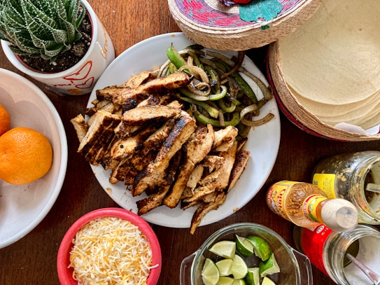 A colorful and inviting spread of food ingredients for making a meal, featuring a plate of grilled chicken strips and saut&eacute;ed onions and peppers, surrounded by elements like shredded cheese, lime wedges, tortillas, orange fruits, various condiments, and a small cactus plant.