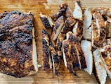Juicy, seasoned, grilled chicken breast sliced and arranged neatly on a wooden cutting board. The chicken is charred to perfection, showcasing a mix of well-cooked edges and tender white interior.