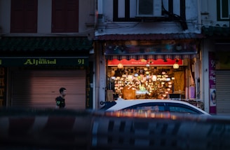 A street scene features a shop with a brightly lit, colorful lantern display inside. The shopfront is decorated with various lamps and ornaments, creating a warm and inviting atmosphere. In the foreground, a white car is parked, and a person is walking past the shop. Other shops with closed shutters are visible under dim lighting, providing a contrast to the vibrant interior of the main shop.