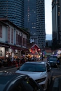 A bustling urban scene featuring a lively street with outdoor dining and people sitting at tables. Traditional low-rise buildings with red roofs and modern high-rise architecture in the background create a contrast. The tall, honeycomb-patterned skyscraper looms above, making the street appear vibrant yet overshadowed by urban development.