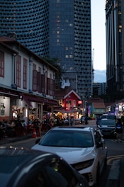 A bustling urban scene featuring a lively street with outdoor dining and people sitting at tables. Traditional low-rise buildings with red roofs and modern high-rise architecture in the background create a contrast. The tall, honeycomb-patterned skyscraper looms above, making the street appear vibrant yet overshadowed by urban development.