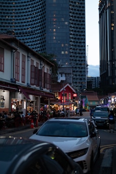 A bustling urban scene featuring a lively street with outdoor dining and people sitting at tables. Traditional low-rise buildings with red roofs and modern high-rise architecture in the background create a contrast. The tall, honeycomb-patterned skyscraper looms above, making the street appear vibrant yet overshadowed by urban development.