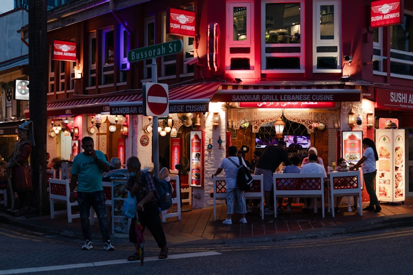 An outdoor dining area with several people sitting at tables under a covered area of a restaurant. The restaurant, called Sanobar Grill Lebanese Cuisine, features bright red signage and decorative hanging lamps. It is located on a street corner with vibrant lighting and a sign for Bussorah Street is visible. Pedestrians are walking by, and the setting is during the evening.