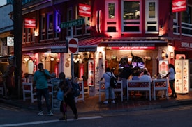 An outdoor dining area with several people sitting at tables under a covered area of a restaurant. The restaurant, called Sanobar Grill Lebanese Cuisine, features bright red signage and decorative hanging lamps. It is located on a street corner with vibrant lighting and a sign for Bussorah Street is visible. Pedestrians are walking by, and the setting is during the evening.