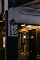 A dimly lit doorway with a sign indicating 'The Gentlemen's Society'. The number 12 is prominently displayed on a white and dark green wall. A vintage-style lamp with a striped pattern is mounted on the wall, casting a warm glow on the entrance. The scene suggests a quaint, possibly exclusive establishment.