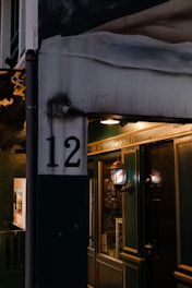 A dimly lit doorway with a sign indicating 'The Gentlemen's Society'. The number 12 is prominently displayed on a white and dark green wall. A vintage-style lamp with a striped pattern is mounted on the wall, casting a warm glow on the entrance. The scene suggests a quaint, possibly exclusive establishment.