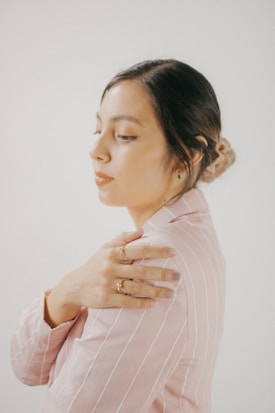 A person with closed eyes wearing a pink and white pinstripe shirt, touching their shoulder gently with their hand adorned with rings. The background is a soft, neutral tone and the lighting is warm, creating a serene atmosphere.