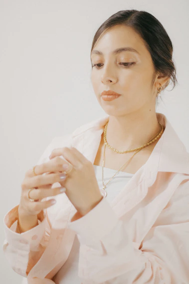 A chic woman wearing delicate gold earrings and a soft pink silk scarf, styled against a beige background.