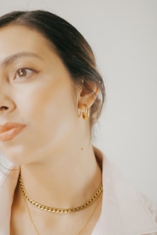Close-up of a woman wearing a simple gold necklace and diamond studs, glowing softly in natural light.