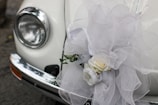 A close-up of a classic car's headlight and bumper, adorned with an elaborate white tulle bow and a floral arrangement containing white roses. The decorations suggest a festive or ceremonial occasion, such as a wedding.