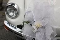 A close-up of a classic car's headlight and bumper, adorned with an elaborate white tulle bow and a floral arrangement containing white roses. The decorations suggest a festive or ceremonial occasion, such as a wedding.