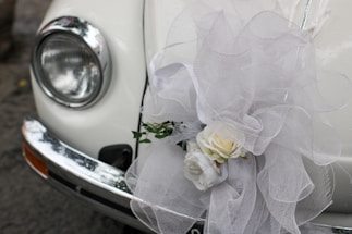 A close-up of a classic car's headlight and bumper, adorned with an elaborate white tulle bow and a floral arrangement containing white roses. The decorations suggest a festive or ceremonial occasion, such as a wedding.