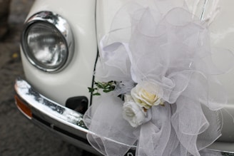 A close-up of a classic car's headlight and bumper, adorned with an elaborate white tulle bow and a floral arrangement containing white roses. The decorations suggest a festive or ceremonial occasion, such as a wedding.