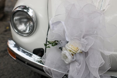 A close-up of a classic car's headlight and bumper, adorned with an elaborate white tulle bow and a floral arrangement containing white roses. The decorations suggest a festive or ceremonial occasion, such as a wedding.