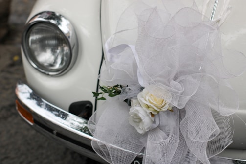 A close-up of a classic car's headlight and bumper, adorned with an elaborate white tulle bow and a floral arrangement containing white roses. The decorations suggest a festive or ceremonial occasion, such as a wedding.