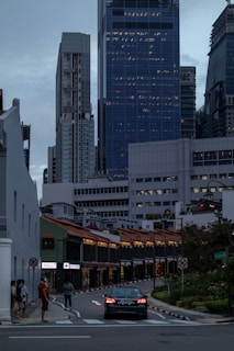 A vibrant city scene featuring modern skyscrapers towering over traditional shophouses. A car is driving on a street lined with illuminated shophouses. People are walking on the sidewalk, and various signs and traffic symbols are visible.