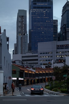A vibrant city scene featuring modern skyscrapers towering over traditional shophouses. A car is driving on a street lined with illuminated shophouses. People are walking on the sidewalk, and various signs and traffic symbols are visible.