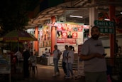 A nighttime street scene outside a cosmetics store called Philms Cosmetic, with bright signage advertising duty-free perfumes. People are walking and socializing around the area. A man in a t-shirt is holding a phone while another group is engaged in conversation near the store. A street sign reading 'Smith St' is visible, along with a colorful umbrella.