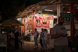 A nighttime street scene outside a cosmetics store called Philms Cosmetic, with bright signage advertising duty-free perfumes. People are walking and socializing around the area. A man in a t-shirt is holding a phone while another group is engaged in conversation near the store. A street sign reading 'Smith St' is visible, along with a colorful umbrella.