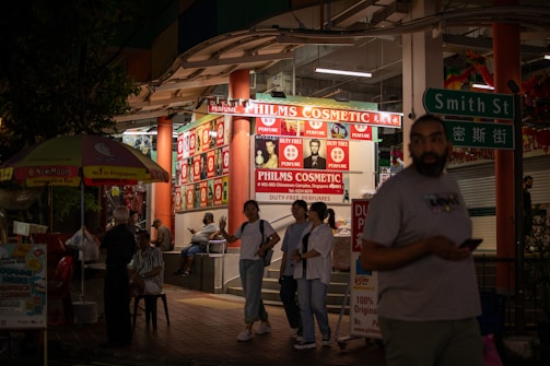 A nighttime street scene outside a cosmetics store called Philms Cosmetic, with bright signage advertising duty-free perfumes. People are walking and socializing around the area. A man in a t-shirt is holding a phone while another group is engaged in conversation near the store. A street sign reading 'Smith St' is visible, along with a colorful umbrella.